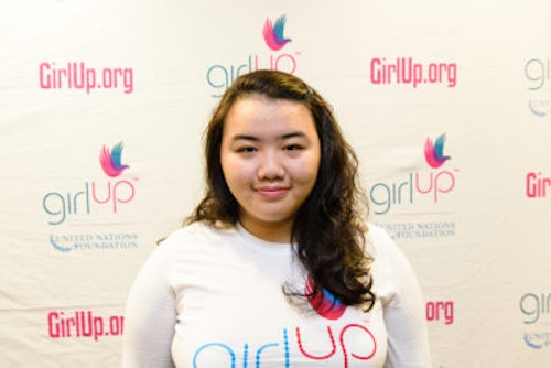 Priscilla Guo_2013-2014 Teen Advisor (close angle headshot, a picture little blurry ) a teen girl wearing her girl up white shirt with her smiley face facing the camera, and background is girlup.org board