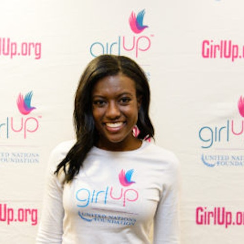 Raven Delk, consultora adolescente de 2013-2014 (foto de perto, um pouco desfocada). Uma adolescente sorridente olhando para a câmera, tendo uma parede com “girlup.org” no plano de fundo. Ela está usando a camiseta toda branca da Girl Up