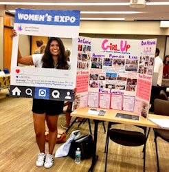 girl up club girl is doing an exhibition with a table of a poster of what Girl Up club do, and she is holding an Instagram sign "women's expo" smile facing to the front