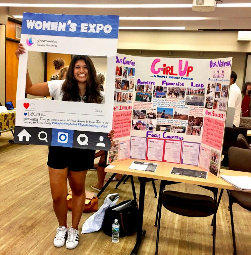 girl up club girl is doing an exhibition with a table of a poster of what Girl Up club do, and she is holding an Instagram sign "women's expo" smile facing to the front