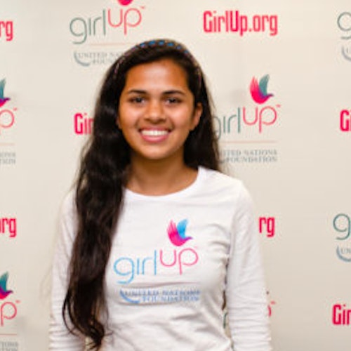 Riya Singh, consultora adolescente de 2012-2013 (foto de perto, um pouco desfocada). Uma adolescente sorridente olhando para a câmera, tendo uma parede com “girlup.org” no plano de fundo. Ela está usando a camiseta toda branca da Girl Up