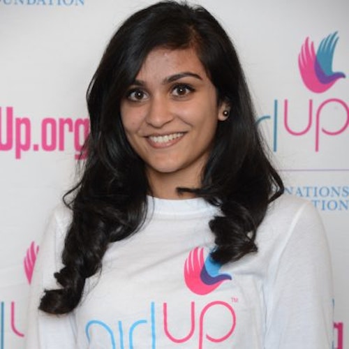 Ruhy Patel_Consejera adolescente 2014-2015 (retrato en primer plano); una adolescente con la camiseta blanca de Girl Up, sonriendo a la cámara, con el cartel de girlup.org de fondo.