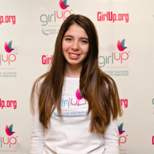 Sarah Gale, copresidente, consultora adolescente de 2013-2014 (foto de perto, um pouco desfocada). Uma adolescente sorridente olhando para a câmera, tendo uma parede com “girlup.org” no plano de fundo. Ela está usando a camiseta toda branca da Girl Up