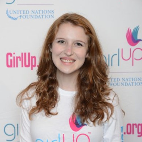 Sarah Gordon, consultora adolescente de 2013-2014 (foto de perto, um pouco desfocada). Uma adolescente sorridente olhando para a câmera, tendo uma parede com “girlup.org” no plano de fundo. Ela está usando a camiseta toda branca da Girl Up