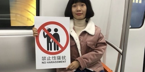 a Chinese girl is sitting down on the metro seat, facing the camera and holding a sign "no harassment."