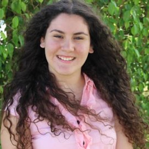 Shayla Zamora, consultora adolescente de 2016-2017 (foto de perto, um pouco desfocada) sorridente olhando para a câmera e tendo o verde das plantas como plano de fundo