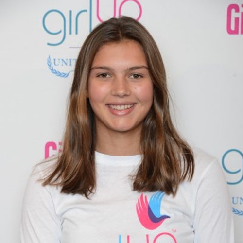 Sydney Baumgardt, consultora adolescente de 2014-2015 (foto de perto). Uma adolescente sorridente olhando para a câmera, tendo uma parede com “girlup.org” no plano de fundo. Ela está usando a camiseta toda branca da Girl Up