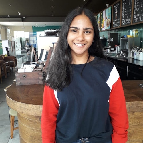 Tanushah Ramadass, consultora adolescente de 2019-2020 (foto de meio-corpo), sorridente olhando para a câmera, tendo uma cafeteria como plano de fundo