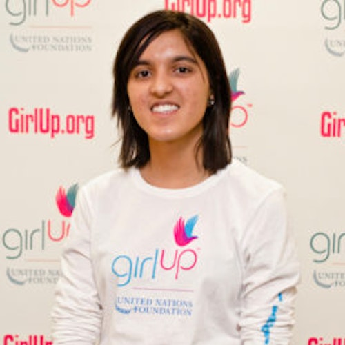 Tanya Devani_家乡： 美国亚拉巴马州伯明翰_2012-2013 届青年顾问（近距离头像照，画面有点模糊），照片中的她穿着白色 Girl Up T 恤，面对镜头微笑，照片背景为 girlup.org 活动展板