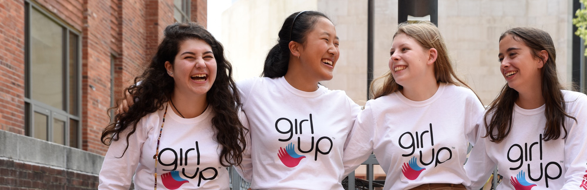 4 名不同种族的 Girl Up 女孩穿着 Girl Up T 恤，手搭在彼此的背上（合影）