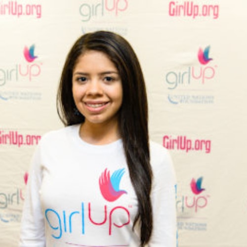 Valeria Hansen_Consejera adolescente 2013-2014 (retrato en primer plano, fotografía un poco borrosa); una adolescente con la camiseta blanca de Girl Up, sonriendo a la cámara, con el cartel de girlup.org de fondo.