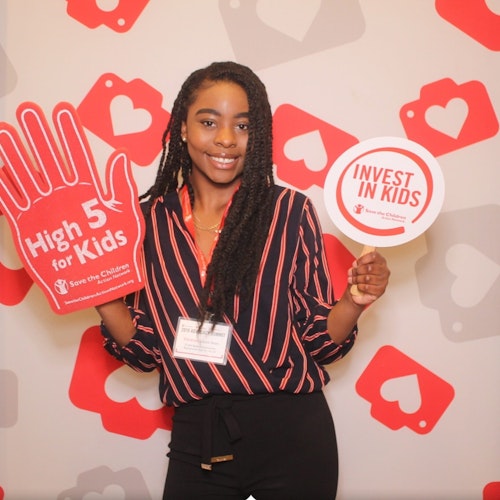 Vanessa Louis-Jean, consultora adolescente de 2019-2020 (foto em plano americano) sorridente olhando para a câmera e segurando as placas “High 5 for kids” (Comemorando com as crianças) e “Invest in kids” (Invista nas crianças)
