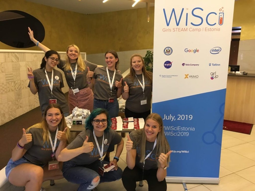 7 girls take group pictures with the WiSci girl STEAM camp standing poster