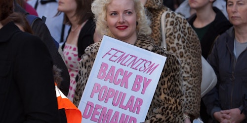 a woman holding a sign "Feminism back by popular demand"