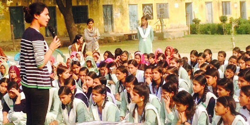 a woman standing and speaking in front of a group of girl students who are sitting on the group listen to her at the school