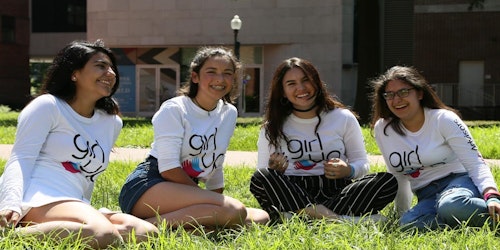 4 فتيات يرتدين قميص المستشارين المراهقين يجلسن على أرضية العشب ويضحكن على الصورة