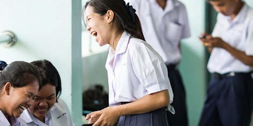 two girls sitting down and looking and their phone and laughing, and one girl standing and laughing holding her phone (two boys in the blurry background also holding their phone)