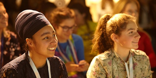 Dos miembros de Girl Up de diferentes orígenes étnicos en Europa sonriendo juntas en un evento mientras escuchan al orador (primer plano).