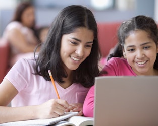 two girls are smiling and writing down notes and sharing a laptop