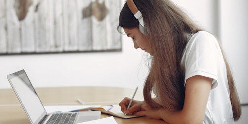 a girl writing notes with her headphone on and a laptop in front of her