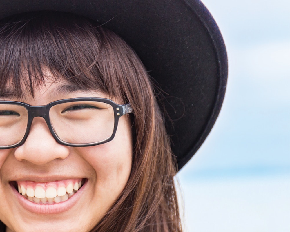 Foto em close muito perto de uma menina sorridente, usando chapéu e óculos