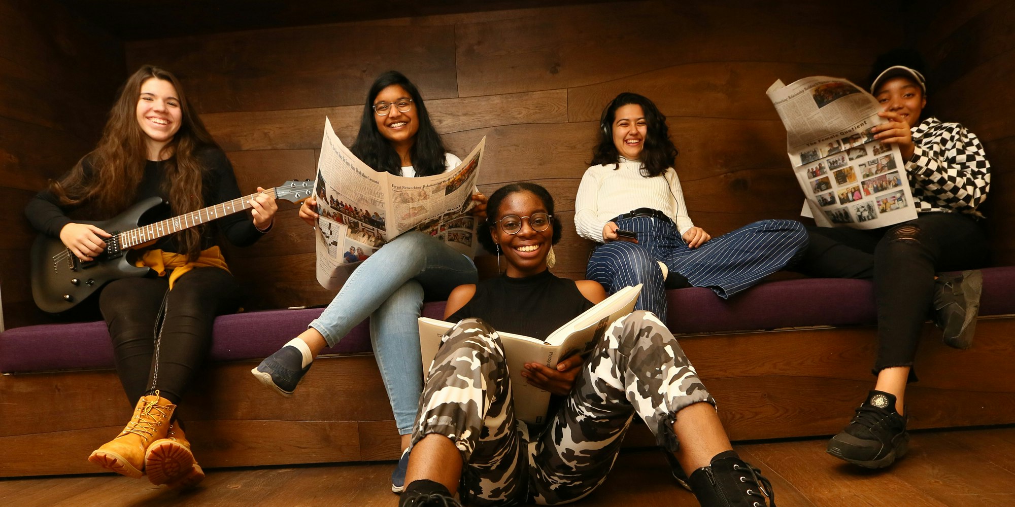 5 chicas de diferentes etnias en la foto sosteniendo libros (sentadas en el piso de enfrente) y sosteniendo la guitarra y el periódico (sentadas en el sofá de atrás)