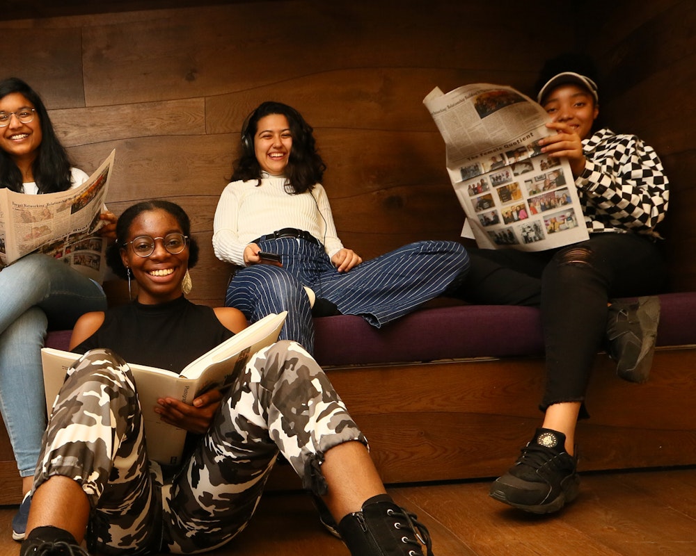 5 名不同种族女孩的合影，坐在前排的女孩拿着书，坐在后面沙发上的女孩拿着吉他和报纸