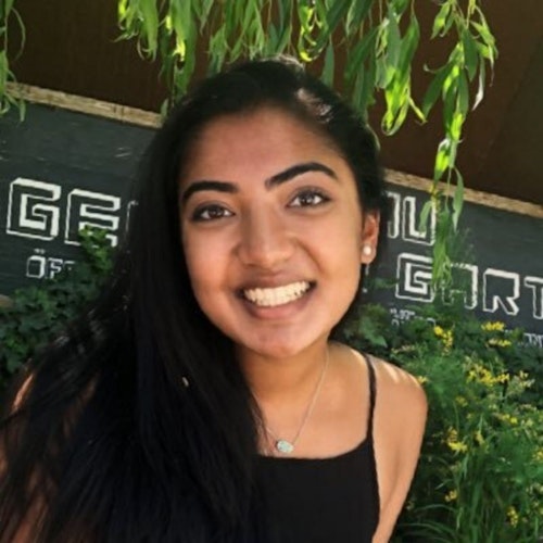 Nehal Jain_ 2015-2016 Asesora de Adolescentes (foto de cabeza de ángulo más amplio con medio cuerpo, foto borrosa) con su cara sonriente mirando a la cámara, el fondo es verde y flores